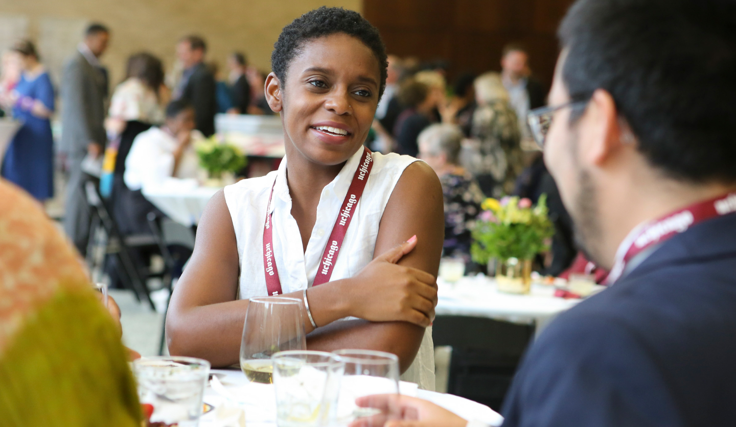 A dark-skinned female-presenting person sits at a banquet table, smiling at a male-presenting person with dark hair.
